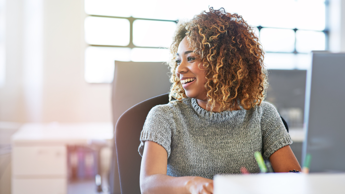 A smiling woman sits at a desk, conveying a sense of positivity and engagement in her work environment.